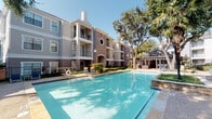 Located on Cedar Springs Road in Dallas, Texas, this community amenity space offers a well-appointed clubhouse and resort-style outdoor area designed for relaxation and socializing. The clubhouse interior features light blue walls, tile flooring, and a retractable glass garage door that opens fully to the pool area, seamlessly blending indoor and outdoor living. Bold orange-cushioned wicker furniture is arranged throughout the space, creating a vibrant and welcoming atmosphere. A built-in wet bar with granite countertops, a microwave, and ample counter space provides convenient entertaining options. A mosaic-tiled accent wall frames a tufted white bench, adding an artistic focal point to the interior lounge area. Recessed lighting and a ceiling fan keep the space comfortable year-round. French doors and large multi-pane windows flood the interior with natural light and offer views of the pool. Outside, a spacious resort-style swimming pool is framed by mature oak and palm trees, with a shaded pergola featuring an oversized U-shaped sectional sofa in matching orange and brown wicker — ideal for group gatherings. The multi-story residential buildings surrounding the courtyard provide an intimate, community-centered setting in the heart of Dallas.