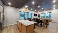 Located at 12000 West Fairview Avenue in Boise, Idaho, this single-floor community amenity space is thoughtfully designed to provide residents with a welcoming and versatile gathering environment. The space is anchored by a full kitchen area, complete with a large center island topped with a striking granite countertop, pendant wicker lighting, wood-toned cabinetry, stainless steel appliances including a refrigerator and built-in oven, and a subway tile backsplash. The kitchen island is surrounded by upholstered bar-height seating, making it ideal for casual dining or social gatherings. Adjacent to the kitchen, a dedicated dining area features dark wood tables and nailhead-trim chairs, with wall-mounted flat-screen televisions positioned for easy viewing. The lounge areas are furnished with comfortable sofas accented by vibrant throw pillows, armchairs, and rustic wood coffee tables, all set atop patterned area rugs over wood-style flooring. Walls throughout are adorned with a bold geometric wallpaper and curated artwork, contributing to a cohesive, modern aesthetic. Large black-framed windows provide ample natural light and offer direct views of the outdoor pool deck and the surrounding residential buildings. The dark ceiling treatment adds a sophisticated contrast to the bright, textured wall design, creating an inviting and well-appointed communal retreat.