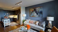 This single-floor space showcases a seamless open-concept design that connects the living area and kitchen into one cohesive, well-appointed environment. The living room serves as the focal point, anchored by a slate-blue accent wall adorned with a large-scale abstract painting that introduces dynamic pops of orange, grey, and white. A tufted grey sofa, layered with patterned and textured throw pillows, sits at the center of the space, complemented by a glass-and-gold coffee table that adds a touch of refinement. Track lighting mounted on the ceiling provides directed illumination, while a floor lamp contributes warm ambient light. Hardwood flooring runs throughout, adding warmth and continuity to the space. The kitchen, visible from the living area, features dark espresso cabinetry with sleek hardware, stainless steel appliances including a full-size refrigerator, and under-cabinet lighting that brightens the workspace. A striped area rug defines the transition between the two zones. Together, the spaces strike a balance between contemporary style and functional living, creating an inviting and visually dynamic environment.