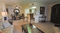 This single-floor residence offers a thoughtfully designed open-concept layout that seamlessly connects the living, dining, and kitchen areas. The living room features plush carpeting, a glass-top coffee table, and a ceiling fan with integrated lighting, contributing to a comfortable and functional atmosphere. Vaulted ceilings add a sense of height and openness to the space, enhancing the overall airy feel. A breakfast bar with a granite-style countertop serves as a natural divider between the living area and the kitchen, which is visible through a bright pass-through opening accented by a skylight above. The kitchen benefits from this natural light, creating a cheerful and inviting cooking environment. The dining area is nestled to the side, furnished with a wooden table and chairs alongside a decorative sideboard topped with a striking circular mirror — adding depth and personality to the room. Warm, neutral tones in soft yellows and beiges flow throughout, creating a cohesive and welcoming ambiance. Wall art and carefully chosen accent pieces lend a curated, homey character to the interior. The layout is practical and well-proportioned, making efficient use of the available space while maintaining a sense of openness and comfort throughout.
