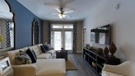 Located at 722 Island Park Drive on Daniel Island, South Carolina, this single-story residence offers a thoughtfully designed living space that blends modern style with comfortable functionality. The main living area serves as the focal point of the home, showcasing rich, dark hardwood-style flooring that grounds the room with warmth and texture. A striking deep blue accent wall adds a bold, contemporary touch, complementing the neutral tones found throughout the space.

Natural light flows generously into the living room through a set of French doors topped with transom windows, which provide direct access to an outdoor balcony—ideal for enjoying the Daniel Island surroundings. Patterned drapes frame the doors, adding visual depth and a layer of elegance. A ceiling fan with an integrated light fixture is centrally positioned overhead, ensuring both comfort and ambient lighting.

The living area is well-appointed with ample space for seating and entertainment, featuring a dedicated media wall with open shelving for storage and display. The overall ambiance of the space is polished yet inviting, with a cohesive color palette and carefully curated details that create a welcoming atmosphere throughout.