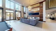 This generously sized single-floor amenity space, identified as The Four at Deerwood, is designed with a contemporary aesthetic that blends sophistication with functionality. The centerpiece of the space is a dramatic lounge area distinguished by soaring double-height ceilings and expansive floor-to-ceiling windows that flood the interior with natural light and offer views of the surrounding landscaping and outdoor areas. A large sectional sofa arrangement anchors the lounge, positioned facing a striking wall-mounted flat-screen television set within a rich wood-paneled feature wall accented by illuminated built-in shelving.

Adjacent to the main lounge, a sleek bar-style counter with white quartz surfaces and pendant lighting serves as a social hub, complemented by a coffee station and workstation nook. The space also includes a dedicated game area featuring a poker table framed by floor-to-ceiling glazing, and a billiards area visible in the layout. Wide-format light tile flooring runs throughout, unifying the open-plan zones. Decorative pendant and chandelier lighting, including a sculptural sputnik-style fixture, add visual interest. A corridor with curated artwork and elevator access completes the thoughtfully appointed space.