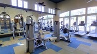 This fitness center offers a comprehensive workout environment designed to accommodate a wide range of exercise routines. The space is characterized by its impressively high ceilings, which create an open, airy ambiance that enhances the overall workout experience. Large windows line the walls, flooding the interior with abundant natural light and offering views of lush outdoor greenery, including palm trees that contribute to a resort-like atmosphere.

The flooring features a distinctive checkered pattern in blue and yellow tones, adding a vibrant visual element to the space. Ceiling fans are mounted throughout, providing additional air circulation to complement the bright, open environment.

The facility is well-stocked with a diverse selection of equipment. Strength training machines, including leg extension and multi-function cable units, are prominently positioned throughout the floor. The cardio section features a row of treadmills, elliptical trainers, and stationary bikes, all positioned near the large windows to allow exercisers to enjoy the outdoor views. Mirrors along the walls further amplify the sense of space while serving a functional purpose for form monitoring during workouts.