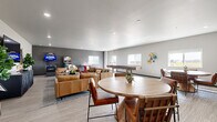 Located at 522 Sweetgrass Dr in Brookings, South Dakota, this single-floor community amenity space offers a well-rounded gathering environment designed for socializing and recreation. The open-concept layout flows seamlessly from the kitchen and dining area into the living and game spaces, all unified by light wood-look flooring and a neutral gray-and-white color palette.

The kitchen is outfitted with dark espresso cabinetry, stainless steel appliances including a full-size refrigerator and dishwasher, a microwave, and a generous granite-topped island with bar seating — ideal for casual dining or entertaining. Adjacent to the kitchen, a round dining table with leather chairs provides an additional gathering spot.

The living area features comfortable tan leather sofas arranged around a coffee table, with a mounted flat-screen TV and recessed lighting throughout. A dedicated game zone includes pinball machines, arcade cabinets, a bubble hockey table, and a full-length shuffleboard table, offering diverse entertainment options. Multiple windows invite natural light throughout the space.

The two restrooms are thoughtfully designed with marble-look tile wainscoting, wood-look tile flooring, bold decorative wallpaper, and accessibility-compliant fixtures, combining style with practicality. Overall, the space presents a modern, well-appointed environment suited for community events and recreation.