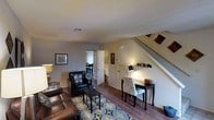 This two-story home offers a thoughtfully arranged layout that balances comfort and functionality across both floors. The main level features an inviting living area anchored by warm hardwood-style flooring, textured ceilings, and neutral wall tones that create a bright and welcoming atmosphere. A plush seating arrangement centers the living room, complemented by a patterned area rug that adds a touch of style and definition to the space. A compact desk area near the staircase provides a convenient workspace, while a "Welcome" display adds a personal touch to the entryway. The staircase leads to the upper floor, offering a natural transition between the two levels. From the living area, sightlines extend through to a dining space and an adjacent room beyond, illustrating the open and connected nature of the floor plan. Natural light filters through the space, enhancing the warm, lived-in ambiance throughout. The overall design reflects a practical yet comfortable home environment, with each area flowing naturally into the next, making the space feel cohesive and well-organized from the moment you step inside.