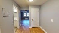 This single-floor space is characterized by warm hardwood flooring that runs throughout, complementing the soft gray-painted walls and crisp white trim and baseboards. A central hallway serves as the spine of the layout, connecting the key areas of the unit. To the left, a bathroom is visible, outfitted with a sink and vanity lighting, finished in neutral tones. Straight ahead, the space opens into a living area distinguished by a double window that draws in natural light, a ceiling fan for air circulation, and a wall-mounted radiator unit indicating radiant heating. The transition between spaces is framed by a wide doorway, lending an open and connected feel to the floor plan. To the right of the hallway, a white paneled interior door with a brass lever handle provides access to an additional room. The flush-mount ceiling light in the hallway and the ceiling fan in the living area provide functional overhead lighting throughout. The overall aesthetic is clean and understated, with a classic residential character reinforced by the wood flooring, traditional trim work, and neutral color palette.