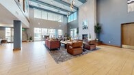 Located at 6504 N 67th Ave in Glendale, Arizona, this single-floor community space offers a thoughtfully designed environment that balances open-concept living with functional amenities. The grand lobby entrance sets an impressive tone with its soaring double-height ceilings, structural columns finished in soft blue-gray tones, warm wood-look tile flooring, and an upper-level mezzanine railing visible overhead.

The expansive living room is the centerpiece of the space, featuring dramatic floor-to-ceiling windows and large sliding glass doors that flood the interior with natural light and provide direct access to an outdoor patio with seating. A striking floor-to-ceiling stone fireplace anchors one wall, complemented by stylish pendant lighting and comfortable seating arrangements with leather and upholstered furnishings atop patterned area rugs.

The kitchen is equally well-appointed, showcasing warm wood cabinetry, dark granite countertops, teal subway tile backsplash, stainless steel appliances, and a large white waterfall-edge island with navy upholstered bar seating. Pendant lights illuminate the workspace, creating an inviting and functional area for gatherings.

Throughout the space, the consistent use of light wood-tone flooring, blue-gray accent walls, and abundant greenery creates a cohesive, welcoming atmosphere suited for communal use.