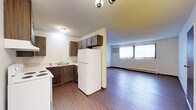 This single-floor space features an open layout that connects the kitchen and living area, creating a functional and cohesive environment. The kitchen is equipped with dark wood cabinetry providing ample storage, a white refrigerator, a double-basin sink, and a white electric range. A textured ceiling with flush-mount lighting illuminates the kitchen workspace efficiently.

The flooring throughout is a dark hardwood-style material that runs seamlessly from the kitchen into the living area, lending visual continuity to the space. The living area is generously proportioned, offering an open and airy feel. A large window fitted with vertical blinds allows natural light to filter in, brightening the room considerably. A baseboard heating unit is positioned beneath the window, and a wall-mounted air conditioning unit provides additional climate control options.

The textured ceiling continues throughout, and the neutral wall tones complement the darker flooring and cabinetry. Overall, the space presents a practical and straightforward layout suited for everyday living.