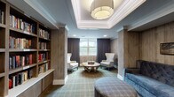 This single-floor library in Germantown, Tennessee, is thoughtfully designed to serve as both a communal gathering space and a quiet retreat for reading and conversation. The layout flows naturally between distinct zones, beginning with a well-appointed lounge area anchored by a tufted gray sofa, plaid upholstered ottomans, and a geometric wooden coffee table. Tall wingback chairs flank the central seating arrangement, providing intimate spots for individual reading. A tray ceiling with elegant drum pendant lighting crowns this area, lending a polished architectural detail overhead.

Floor-to-ceiling built-in bookshelves line one wall, stocked with a diverse collection of titles and providing a visual centerpiece that reinforces the library's purpose. Warm wood-tone wall paneling throughout the space contributes to a cozy yet refined ambiance, complemented by soft, patterned carpeting in muted greens and tans.

Large windows dressed with rich, dark curtain panels allow natural light to filter in while offering views of an outdoor courtyard and surrounding greenery. A semi-private nook at one end of the room features a round table surrounded by teal upholstered chairs, ideal for small group discussions or games. Curated wall art and subtle décor details complete the sophisticated, welcoming atmosphere of this versatile library space.