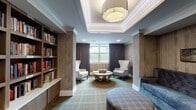 This single-floor library in Germantown, Tennessee, is thoughtfully designed to serve as both a communal gathering space and a quiet retreat for reading and conversation. The layout flows naturally between distinct zones, beginning with a well-appointed lounge area anchored by a tufted gray sofa, plaid upholstered ottomans, and a geometric wooden coffee table. Tall wingback chairs flank the central seating arrangement, providing intimate spots for individual reading. A tray ceiling with elegant drum pendant lighting crowns this area, lending a polished architectural detail overhead.

Floor-to-ceiling built-in bookshelves line one wall, stocked with a diverse collection of titles and providing a visual centerpiece that reinforces the library's purpose. Warm wood-tone wall paneling throughout the space contributes to a cozy yet refined ambiance, complemented by soft, patterned carpeting in muted greens and tans.

Large windows dressed with rich, dark curtain panels allow natural light to filter in while offering views of an outdoor courtyard and surrounding greenery. A semi-private nook at one end of the room features a round table surrounded by teal upholstered chairs, ideal for small group discussions or games. Curated wall art and subtle décor details complete the sophisticated, welcoming atmosphere of this versatile library space.