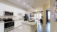 This stylish single-floor residence showcases a thoughtfully designed open-concept layout that seamlessly connects the kitchen and living areas. The kitchen is a standout feature, boasting high-gloss white cabinetry that extends to the ceiling, a classic white subway tile backsplash, and a generous island with a light quartz countertop — ideal for meal prep and casual dining. Stainless steel appliances, including a French door refrigerator, built-in microwave, and range, add a professional touch to the space. Elegant pendant lighting with crystal accents hangs above the island, complementing the recessed lighting throughout. Light hardwood-style flooring runs continuously from the kitchen into the living area, enhancing the sense of flow and openness. The living space is bathed in natural light streaming through floor-to-ceiling windows dressed with patterned curtains, offering a bright and inviting ambiance. Neutral wall tones and contemporary finishes throughout create a clean, modern aesthetic that is both functional and visually appealing, making this residence a welcoming and well-appointed home.