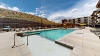 The outdoor amenity area at 186 E Future Way is a thoughtfully designed resort-style space set against a backdrop of natural desert hillsides. At its centerpiece sits a large, sparkling swimming pool with entry steps and surrounding pool deck, generously furnished with sling-style chaise lounges, side tables, and vibrant yellow market umbrellas that add a lively pop of color throughout the space. Adjacent to the pool, a hot tub provides an additional relaxation option, while a dedicated cornhole court offers casual outdoor entertainment. The surrounding hardscape is clean and expansive, providing ample room for lounging and socializing. A compact pool house building finished in natural stone and wood siding serves the area and includes access to restroom facilities. The space is enclosed by sleek black iron fencing, with landscaped borders featuring drought-tolerant grasses, ornamental shrubs, and young trees that soften the hardscape. Curving pathways connect various zones of the amenity area and lead to gated entries. The multi-story residential building, clad in a modern blend of stone veneer and horizontal wood-tone siding with private balconies, frames the space and creates a cohesive, community-centered environment open to the sky and surrounding natural scenery.