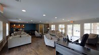 This single-story space is thoughtfully designed to serve as a functional and inviting community or leasing hub. The open-plan layout seamlessly connects multiple zones, including a well-appointed lounge area, a dining nook, and a front-desk or office station. The lounge is furnished with neutral-toned sofas and elegantly patterned accent chairs arranged around area rugs, creating a comfortable and welcoming atmosphere for visitors. A bold teal accent wall anchors the dining area at the rear, providing a striking contrast to the warm, neutral tones found throughout the rest of the space. Wood-look plank flooring runs consistently throughout, adding warmth and a cohesive design aesthetic. Recessed ceiling lighting is evenly distributed across the smooth, painted ceiling, ensuring bright and consistent illumination. Large windows and glass patio doors along the far wall flood the interior with natural light while offering views of the surrounding outdoor landscape. A front desk or workspace with modern, dark-finished countertops is positioned near the entrance, facilitating a professional point of contact. Decorative floor lamps and framed artwork further enhance the refined yet approachable ambiance of this versatile, single-level space.