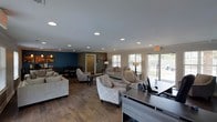 This single-story space is thoughtfully designed to serve as a functional and inviting community or leasing hub. The open-plan layout seamlessly connects multiple zones, including a well-appointed lounge area, a dining nook, and a front-desk or office station. The lounge is furnished with neutral-toned sofas and elegantly patterned accent chairs arranged around area rugs, creating a comfortable and welcoming atmosphere for visitors. A bold teal accent wall anchors the dining area at the rear, providing a striking contrast to the warm, neutral tones found throughout the rest of the space. Wood-look plank flooring runs consistently throughout, adding warmth and a cohesive design aesthetic. Recessed ceiling lighting is evenly distributed across the smooth, painted ceiling, ensuring bright and consistent illumination. Large windows and glass patio doors along the far wall flood the interior with natural light while offering views of the surrounding outdoor landscape. A front desk or workspace with modern, dark-finished countertops is positioned near the entrance, facilitating a professional point of contact. Decorative floor lamps and framed artwork further enhance the refined yet approachable ambiance of this versatile, single-level space.