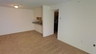 This single-floor residential unit offers a straightforward, functional layout designed for comfortable everyday living. The living area is open and airy, featuring wall-to-wall beige carpeting that adds warmth and continuity throughout the space. Neutral, light-colored walls keep the interior feeling bright and clean, providing a versatile backdrop for a variety of décor styles.

A defining architectural feature is the half-wall pass-through that connects the living area to the kitchen, creating a sense of openness while maintaining a visual distinction between the two spaces. The kitchen is equipped with wood-toned cabinetry, a single-basin sink, and countertops with a granite-style finish, offering a practical and organized cooking environment. A thermostat is conveniently mounted on the wall near the kitchen entry, indicating climate control throughout the unit.

An adjacent doorway leads to a separate area — likely a bedroom or additional room — partially visible and hinting at a private retreat beyond the main living space. The overall ambiance is clean, neutral, and move-in ready, making it well-suited for those seeking a functional and low-maintenance living environment.