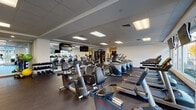Located at 109 Ashe Ave in Raleigh, NC, this single-floor fitness center offers a comprehensive workout environment designed to accommodate a wide range of exercise routines. The space is thoughtfully organized into distinct functional zones, each outfitted with professional-grade Life Fitness equipment throughout.

The cardio area is generously stocked with multiple treadmills, elliptical trainers, and recumbent and upright stationary bikes, all arranged in orderly rows across rubber-floored surfaces that provide cushioning and durability. Large windows along one wall allow abundant natural light to fill the space while offering views of the surrounding streetscape, creating an open and motivating atmosphere.

The strength and free-weight zone features a well-stocked dumbbell rack, weight benches, stability balls, resistance bands, and cable machines. Full-length mirrors line sections of the wall, visually expanding the space and allowing users to monitor their form during exercise. A vibrant green and white graphic mural adds a dynamic visual element to one wall, contributing to the energetic ambiance.

Multiple wall-mounted flat-screen televisions are positioned throughout the facility for entertainment during workouts. The dropped ceiling with recessed panel lighting ensures the space remains consistently well-lit. Overall, the fitness center presents a clean, modern, and fully functional environment.