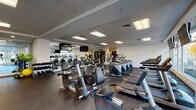 Located at 109 Ashe Ave in Raleigh, NC, this single-floor fitness center offers a comprehensive workout environment designed to accommodate a wide range of exercise routines. The space is thoughtfully organized into distinct functional zones, each outfitted with professional-grade Life Fitness equipment throughout.

The cardio area is generously stocked with multiple treadmills, elliptical trainers, and recumbent and upright stationary bikes, all arranged in orderly rows across rubber-floored surfaces that provide cushioning and durability. Large windows along one wall allow abundant natural light to fill the space while offering views of the surrounding streetscape, creating an open and motivating atmosphere.

The strength and free-weight zone features a well-stocked dumbbell rack, weight benches, stability balls, resistance bands, and cable machines. Full-length mirrors line sections of the wall, visually expanding the space and allowing users to monitor their form during exercise. A vibrant green and white graphic mural adds a dynamic visual element to one wall, contributing to the energetic ambiance.

Multiple wall-mounted flat-screen televisions are positioned throughout the facility for entertainment during workouts. The dropped ceiling with recessed panel lighting ensures the space remains consistently well-lit. Overall, the fitness center presents a clean, modern, and fully functional environment.