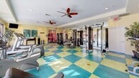 Located in La Quinta, California, this well-equipped fitness center offers a bright and inviting environment for a complete workout experience. The space is defined by its distinctive checkered teal and yellow-green flooring, which adds a vibrant, energetic character to the room. Large glass French doors line one wall, flooding the interior with abundant natural light and providing direct views of the outdoor courtyard and surrounding property, creating an open and airy atmosphere.

The facility is comprehensively outfitted with a full array of cardio equipment, including multiple treadmills, elliptical trainers, and a stationary bike, all positioned to take advantage of the natural light and outdoor views. The strength training area features a multi-station cable machine, a weight bench, and a full dumbbell rack, catering to a wide range of exercise routines. Full-length mirrors along one wall visually expand the space and provide practical utility for form-checking during workouts.

Recessed lighting and dual ceiling fans with light fixtures ensure consistent airflow and comfortable conditions throughout the room. Warm yellow walls contribute to the space's welcoming ambiance, while decorative artwork and potted plants add subtle finishing touches. The overall design balances functionality with an uplifting aesthetic suited to the sunny Southern California setting.