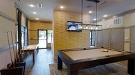 This single-floor recreation space is thoughtfully designed to serve as a social hub for games and leisure. The room's centerpiece is a sleek, dark-wood pool table positioned beneath a contemporary rectangular pendant light fixture, adding a polished, modern aesthetic to the space. A textured, undulating accent wall in warm sandy tones serves as a striking architectural focal point, anchoring a wall-mounted flat-screen television that makes the area ideal for entertaining.

Adjacent to the pool table, a shuffleboard table provides additional gaming options, catering to a variety of recreational interests. The flooring features wood-look tile laid in a clean, linear pattern, lending a sophisticated yet durable finish throughout.

Recessed lighting in the ceiling provides consistent, well-distributed illumination across the space, complemented by natural light streaming in through large windows and a glass door that opens to an outdoor patio area. The outdoor space, visible through the glazing, features umbrella-shaded seating, extending the social environment beyond the interior.

The overall ambiance is contemporary and inviting, combining stylish finishes with practical amenities. The open layout encourages movement and interaction, making this a versatile space well-suited for casual gatherings and recreational activities alike.