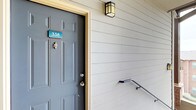 This single-floor, 2-bedroom, 2-bathroom residence offers a thoughtfully designed layout that maximizes functionality and comfort. The entry is marked by a charming craftsman-style wall sconce and a bold gray paneled front door, creating an inviting first impression complemented by horizontal lap siding along the exterior facade.

Inside, the open living area features neutral carpet flooring, crisp white walls, and dark wood trim accents that frame doorways and baseboards throughout, providing clean visual contrast. A hallway extends toward the bedrooms, lined with dark-trimmed doors and connecting the living space to the private quarters. Overhead ventilation is integrated seamlessly into the ceiling, contributing to the unit's practical design.

The kitchen is partially visible and features granite-style countertops, offering a functional workspace that complements the overall aesthetic. Natural light flows through windows at the end of the hallway, keeping the interior bright.

One of the standout features of this residence is the private balcony, which offers expansive views of the surrounding brick building community, lush treetops, and open sky—providing a tranquil outdoor retreat with a scenic, elevated perspective.
