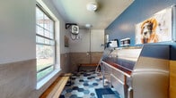 Located at 9170 Crowne Springs View in Colorado Springs, CO, this purpose-built pet wash station is a thoughtfully designed, single-room facility tailored for pet grooming needs. The space is immediately distinguished by its striking hexagonal tile flooring, which blends shades of blue, gray, and tan to create a visually dynamic yet practical surface. The lower half of the walls is clad in neutral rectangular tile, while the upper portion is painted in a bold slate blue, lending the room a clean, modern aesthetic. At the center of the space sits a large, deep stainless steel grooming tub equipped with a flexible handheld spray hose, providing a fully functional bathing setup. A wall-mounted electric heater and a mini-split HVAC unit ensure year-round climate comfort for both pets and their owners. Plumbing infrastructure, including an on-demand water system, is visibly integrated along the blue accent wall. Wooden slatted benches offer convenient seating along the perimeter. Large windows allow generous natural light to fill the room while providing views of the surrounding outdoor greenery. Charming framed dog-themed artwork adds a welcoming, playful touch to the walls. The space is accessed through a dark-paneled entry door with glass insets and is equipped with a wall-mounted leash hook and waste receptacle for added convenience.