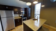 This single-floor space showcases a well-appointed kitchen designed with both functionality and style in mind. The kitchen is anchored by rich, dark wood cabinetry that extends to the ceiling, providing generous storage throughout. Stainless steel appliances, including a refrigerator, electric range with oven, and built-in microwave, lend a sleek, contemporary feel to the space.

A standout feature is the central island, topped with light-toned granite countertops that contrast elegantly with the dark cabinetry. The island incorporates a stainless steel sink with a modern gooseneck faucet, making it a practical hub for meal preparation. Matching granite countertops line the perimeter of the kitchen, reinforcing a cohesive aesthetic.

Natural light filters in through a window fitted with horizontal blinds, brightening the space alongside stylish pendant and drum ceiling fixtures. The light hardwood flooring adds warmth and complements the overall color palette. The exposed ceiling structure contributes an industrial character, giving the kitchen a loft-like ambiance that balances modern finishes with raw architectural detail.