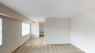 This single-floor unit is laid out with a practical, straightforward design finished in neutral tones throughout. The living area is generously sized, featuring wall-to-wall carpeting and multiple large windows fitted with horizontal blinds, allowing ample natural light to fill the space. A transition to tile flooring near the entryway provides a clear delineation between the entry and living zones. Flush-mount ceiling lighting keeps the space well-illuminated.

A separate room, visible through an open doorway, functions as a bedroom and includes carpeted flooring, a window overlooking a green outdoor view, and a bifold closet door offering built-in storage. The clean white walls and neutral palette carry consistently through each room.

The kitchen features warm-toned wood cabinetry with gold hardware on both upper and lower units, light-colored laminate countertops, and a full-size white refrigerator. The layout is compact and efficient, maximizing available storage and counter space.

The bathroom includes a full-size soaking tub with a showerhead and chrome fixtures, enclosed by taupe square ceramic tile extending up the walls. A chrome towel bar and shower curtain rod complete the functional setup. Overall, the unit presents a clean, move-in ready environment with consistent finishes throughout.