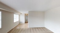 This single-floor unit is laid out with a practical, straightforward design finished in neutral tones throughout. The living area is generously sized, featuring wall-to-wall carpeting and multiple large windows fitted with horizontal blinds, allowing ample natural light to fill the space. A transition to tile flooring near the entryway provides a clear delineation between the entry and living zones. Flush-mount ceiling lighting keeps the space well-illuminated.

A separate room, visible through an open doorway, functions as a bedroom and includes carpeted flooring, a window overlooking a green outdoor view, and a bifold closet door offering built-in storage. The clean white walls and neutral palette carry consistently through each room.

The kitchen features warm-toned wood cabinetry with gold hardware on both upper and lower units, light-colored laminate countertops, and a full-size white refrigerator. The layout is compact and efficient, maximizing available storage and counter space.

The bathroom includes a full-size soaking tub with a showerhead and chrome fixtures, enclosed by taupe square ceramic tile extending up the walls. A chrome towel bar and shower curtain rod complete the functional setup. Overall, the unit presents a clean, move-in ready environment with consistent finishes throughout.