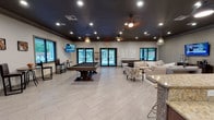 Located at 6721 Washington Ave in Ocean Springs, Mississippi, this single-floor recreation room is thoughtfully designed for both socializing and relaxation. The open-concept layout is anchored by a striking full-size billiards table at its center, positioned beneath elegant geometric pendant lighting and set against light-toned large-format tile flooring that runs throughout the space.

The main gathering area features a comfortable lounge with plush cream sofas, a glass-and-gold coffee table, and a wall-mounted flat-screen TV framed by textured accent wallcovering — ideal for casual viewing or group conversations. Additional seating is provided by bistro-style tables and chairs positioned near the windows, which allow natural light to fill the space and offer views of the outdoor area beyond the glass doors.

A fully equipped kitchen area serves as a functional hub for events and gatherings. Rich dark cabinetry pairs with granite countertops and a slate-blue tile backsplash, complemented by stainless steel appliances including a refrigerator and microwave. A wraparound bar counter with mosaic tile detailing and rustic wood stools provides ample space for dining and entertaining. The dark ceiling treatment throughout ties the design together, creating a sophisticated and inviting atmosphere suited for community recreation and private events.