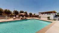 This outdoor pool facility in Moreno Valley, California, offers a well-appointed recreational retreat set against a striking backdrop of rugged mountain terrain and clear Southern California skies. The centerpiece of the space is a generously sized swimming pool with pristine, crystal-clear water, complemented by a separate spa for relaxation. The surrounding pool deck is finished in smooth, light-toned concrete, providing ample space for lounging and entertaining. Multiple chaise lounge chairs with shading umbrellas are positioned throughout the deck, offering comfortable areas to relax in the sun or shade. A covered pergola-style pavilion on one side of the deck houses an outdoor BBQ station, making the space well-suited for social gatherings. The pool area is fully enclosed by a black wrought-iron safety fence. Anchoring the space is a Spanish Colonial-style clubhouse building, distinguished by its terracotta red tile roof, arched entryways, white stucco exterior, and large multi-pane windows that add architectural character to the setting. The combination of the scenic mountain views, resort-style amenities, and warm California climate creates an inviting and functional outdoor community space.