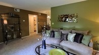 This single-floor residence showcases a thoughtfully designed open-concept layout connecting the living and dining areas. The living room is anchored by a plush, light-toned sectional sofa adorned with decorative throw pillows in earthy tones, positioned against a bold sage green accent wall featuring an abstract metallic wall sculpture. A complementary deep brown accent wall adds warmth and visual contrast to the space. Plush wall-to-wall carpeting runs throughout the living area, contributing to a cozy ambiance. A round glass-and-metal coffee table serves as a central focal point, styled with a sculptural vase of white florals and a chartreuse decorative plate. The adjacent dining area is defined by a pendant light fixture hanging above a dark dining table and chairs, seamlessly continuing the earthy, contemporary color palette. A hallway visible from the main living space leads to what appears to be a bathroom, offering a glimpse of additional functional areas within the home. The overall design aesthetic blends rich, saturated tones with contemporary furnishings to create an inviting and cohesive living environment.