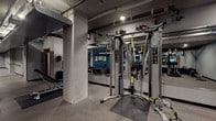 Located at 624 Yale Avenue North in Seattle's South Lake Union neighborhood, this lower-level amenity space offers a comprehensive fitness and bike storage facility designed to serve an active urban lifestyle. The fitness center is well-appointed with a wide range of equipment, including cardio machines such as treadmills and rowing machines, a multi-functional cable machine with TRX suspension trainers, free weights, a weight bench, and a stationary bike. Full-length mirrors line the walls, enhancing the sense of openness throughout the workout area, while wall-mounted displays provide entertainment or guided fitness programming. Rubber flooring throughout the gym ensures durability and safety. A dedicated open area with a yoga mat provides space for stretching or floor exercises. The exposed concrete columns and overhead ductwork give the space an industrial character, complemented by recessed lighting that keeps the environment well-lit. Adjacent to the gym, a spacious bike storage room features wall-mounted bike racks accommodating numerous bicycles, along with a dedicated bike repair station equipped with sturdy workbenches and mounted tools. Restrooms with tiled finishes are conveniently accessible from the fitness area, completing this practical and functional amenity space.
