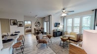 Located at 1626 N Ellison Dr in the Estraya Hills community of San Antonio, TX, this single-floor residence offers a thoughtfully designed open-concept layout with contemporary finishes throughout. The living area is bright and welcoming, with large windows dressed in elegant grommet curtains that invite abundant natural light and offer views of the surrounding community. Light wood-look flooring, a ceiling fan, and a wall-mounted TV anchor the space, while the open flow connects seamlessly to the dining area, which features a sliding glass door providing outdoor access. The kitchen is well-appointed with dark cabinetry, stainless steel appliances, a granite-topped island with bar seating, and track lighting overhead. Two bedrooms are carpeted for comfort and each features ceiling fans, large windows with blinds and curtains, and ample space. The primary bedroom is serene and generously proportioned, while the second bedroom offers equally comfortable accommodations. Both bathrooms are finished with gray tile surrounds, soaking tubs, dark wood cabinetry, and granite countertops, providing a clean, modern aesthetic. Throughout the residence, a neutral color palette and cohesive design elements create a warm, contemporary ambiance ideal for everyday living.