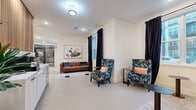 Located at 9821 Research Drive in Charlotte, NC, this well-appointed single-floor amenity space offers a thoughtfully designed layout across multiple functional areas. The two living room lounge areas are bright and welcoming, bathed in natural light streaming through large windows dressed with dramatic floor-to-ceiling black curtains. These spaces are furnished with a rich brown tufted leather sofa, vibrant floral-patterned wingback chairs, and a mix of round accent tables, all set atop textured area rugs against light, neutral-toned walls and smooth concrete-style flooring. Curated wall art adds a refined finishing touch to the relaxed, conversational atmosphere. The dedicated office area provides a more focused environment, seamlessly accessible from the main lounge. The standout feature of the space is the conference room, distinguished by a striking floor-to-ceiling black mural adorned with intricate white scrollwork and a prominent "Queen City" crown motif — a bold nod to Charlotte's nickname. A solid wood conference table surrounded by upholstered gray chairs sits beneath a modern globe chandelier, while a large window offers views of the surrounding neighborhood. Together, these spaces create a versatile, design-forward environment ideal for both collaboration and relaxation.
