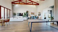 This single-floor community clubhouse is defined by its dramatic vaulted ceiling adorned with bold, red-stained exposed wood trusses, creating an impressive architectural focal point throughout the main gathering space. Wood-look tile flooring spans the entire interior, lending warmth and visual continuity across all areas.

The expansive main hall is thoughtfully organized into distinct social zones. A lounge area features plush white sofas, a wall-mounted flat-screen TV, and decorative indoor olive trees set in large ceramic urns, while pendant and recessed lighting provide a well-lit, welcoming atmosphere. A ping-pong table anchors the recreation area, and a bar-height counter with teal stools offers a casual seating arrangement for socializing or working.

A separate seating nook with floor-to-ceiling windows and arched accents floods the space with natural light, framing views of palm trees and the surrounding outdoor landscape. Tan leather armchairs and elegant drapery give this corner an intimate, refined feel.

A compact kitchenette is equipped with stainless steel appliances, a dishwasher, a mini refrigerator, and clean white countertops accented by curated artwork. A dining area near the arched glass entry doors features a round wood table with leather chairs and lush tropical plants.

Expansive multi-panel glass doors open directly to a covered outdoor patio with arched columns, extending the living and entertaining space seamlessly into the outdoors.