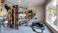 This fitness center is a comprehensively outfitted facility designed to support a wide range of workout routines. The space is finished with neutral, textured wall coverings, crown molding, and blue-toned carpet flooring throughout, creating a clean and polished aesthetic. Recessed lighting and a ceiling fan ensure the environment remains comfortable during use.

The cardio zone is positioned along a wall lined with large windows, allowing abundant natural light to pour in alongside views of surrounding greenery and an outdoor pool area. This section houses multiple treadmills, elliptical trainers, and a stationary bike, each accompanied by wall-mounted flat-screen televisions.

A dedicated strength training area features a robust functional training rig in black and orange, stocked with medicine balls and accessories. A full dumbbell rack and additional resistance equipment round out the free weights section. Floor-to-ceiling mirrors line one wall, visually expanding the space and allowing users to monitor their form.

An adjacent stretch and warm-up zone offers open floor space, while coat hooks and a waste bin near the pool-facing windows add practical convenience. The fitness center is accessible through a clearly marked glass-door entrance.