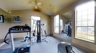 This fitness center offers a practical and comfortable environment for a full workout experience. The room features vaulted ceilings accented by a ceiling fan, which promotes air circulation and adds an open, airy feel to the space. Warm, neutral-toned walls complement the durable, cushioned flooring designed to withstand the demands of regular exercise.

Natural light streams in through multiple windows positioned along the walls, creating a bright and inviting atmosphere throughout the day. A large wall-mounted mirror spans a significant portion of one wall, enhancing the sense of space while allowing users to monitor their form during workouts.

The fitness center is outfitted with a multi-station cable and strength training machine, ideal for a wide range of resistance exercises targeting various muscle groups. Cardio equipment, including a treadmill and an additional cardio machine, rounds out the offerings and supports both warm-up routines and sustained aerobic training sessions. A set of free weights is also visible, providing further versatility for strength training.

Overall, the fitness center is thoughtfully laid out to maximize usability within a compact footprint, making it a convenient and functional amenity for residents or guests seeking an on-site workout facility.