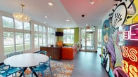 Located at 916 N College Ave in Bloomington, Indiana, this single-floor community space is designed with a bold, energetic aesthetic that makes a strong visual impression from the moment you enter. The layout is open and expansive, encompassing distinct functional zones that flow seamlessly into one another.

The main lounge area is anchored by a vibrant orange sectional sofa and matching armchair, arranged atop a swirling teal area rug and oriented toward a large wall-mounted flat-screen television. Floor-to-ceiling windows line the walls, inviting an abundance of natural light and offering clear views of the surrounding greenery and streetscape outside.

A front reception or check-in desk with warm wood cabinetry provides a welcoming focal point near the entrance, which is framed by a large, eye-catching pop-art mural. A secondary lounge area features a gray sofa, red accent chairs, a bold red-and-white graphic wall mural, and a curated arrangement of framed artwork, adding a gallery-like quality to the space.

A casual dining area rounds out the layout, furnished with bistro-style high-top tables and sleek modern chairs beneath tall, narrow windows and a sculptural chandelier. Throughout the space, a mix of tile and hard flooring, recessed lighting, and vibrant accent walls in lime green, orange, and teal create a cohesive, contemporary, and energetic atmosphere well-suited for social gathering and community engagement.