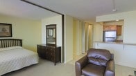 This single-floor residence offers an open, efficient layout that seamlessly integrates the living, sleeping, and kitchen areas into one cohesive space. The sleeping area is defined by a partial room divider, creating a sense of separation while maintaining the open flow of the floor plan. Soft, neutral-toned walls and light carpeting throughout lend a warm and understated ambiance to the interior.

The living area accommodates comfortable seating, as seen with the rich brown leather armchair positioned to enjoy the open layout. Adjacent to the sleeping area, a set of bifold closet doors provides ample storage, while a secondary closet or utility space is visible along the hallway corridor.

The kitchenette, visible to the right, features warm wood cabinetry and a countertop workspace, offering the essential amenities for everyday meal preparation. A window beside the kitchen area allows natural light to filter into the space.

The textured ceiling adds a classic residential character throughout. Overall, the space is thoughtfully arranged to maximize functionality within a compact footprint, making it a practical and comfortable living environment suited for individuals seeking an all-in-one residential layout.