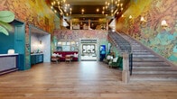 Located at 425 Devon Bryan St. in Columbus, OH, this two-floor clubhouse for Kempton Luxury Living is a thoughtfully designed amenity space that blends eclectic artistry with refined comfort across a generous footprint. The welcoming entrance greets visitors with floor-to-ceiling windows framed in deep green millwork, flooding the space with natural light and offering views of the surrounding community. Warm hardwood floors carry throughout the main level, anchoring a series of distinct gathering spaces. A vibrant lounge area showcases bold, large-scale murals and curated gallery walls, complemented by a built-in café bar with teal cabinetry, creating an energetic yet inviting social hub. A more relaxed seating area features richly upholstered leather and velvet furniture set against sophisticated wainscoting and eclectic portrait artwork. The upper floor houses a private screening room with a large projection screen, ornate globe chandeliers, plush lounge seating, a popcorn machine, and dramatic patterned carpeting. Additional spaces include intimate high-top seating nooks bathed in natural light, a cozy lounge with patterned carpets and leather armchairs, and a dedicated indoor golf simulator room equipped with a TrackMan system and full turf bay, offering a premium recreational experience year-round.
