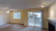 This single-floor residential unit offers a welcoming and functional layout designed for comfortable everyday living. The living area is the heart of the space, characterized by soft wall-to-wall carpeting and warm yellow-toned walls that create a cozy yet airy atmosphere. Natural light fills the room generously, courtesy of a large picture window and a full-width sliding glass door, both of which frame views of mature evergreen trees and lush surrounding greenery. The sliding glass door opens directly onto a private balcony enclosed by a classic white railing, providing an inviting outdoor extension of the living space. The textured ceiling adds a traditional architectural touch consistent throughout the unit. Ceiling-mounted lighting fixtures provide additional illumination when needed. The open layout allows for flexible furniture arrangements, making the living area suitable for a variety of lifestyle needs. The overall ambiance of the space is calm and residential, with an emphasis on natural light, outdoor connectivity, and a serene, tree-lined outlook that brings a sense of nature indoors.