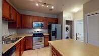 This single-floor unit is thoughtfully laid out to maximize functionality across its living spaces. The kitchen serves as a central highlight, outfitted with rich cherry wood cabinetry offering generous storage both above and below the counters. Stainless steel appliances include an electric range, over-the-range microwave, dishwasher, and a side-by-side refrigerator with an in-door ice and water dispenser. Light-toned countertops complement the warm cabinetry, while large-format tile flooring adds durability and easy maintenance. A kitchen island provides additional prep and storage space. Conveniently situated just off the kitchen is a dedicated laundry alcove housing a stacked washer and dryer unit. The bedroom is finished with plush wall-to-wall carpeting in a neutral tone and features a window with blinds that allows natural light into the space. A ceiling fan is also present in the living area, contributing to overall comfort. The bathroom is clean and functional, featuring a tub and shower combination with white subway tile surround, a single-sink vanity with a wood cabinet base, tile flooring, and neutral gray walls that create a calm, understated aesthetic throughout.
