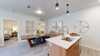 This thoughtfully designed single-floor residence combines modern aesthetics with functional living. The open-concept kitchen is a standout feature, outfitted with stainless steel appliances including a gas range, microwave, and refrigerator, complemented by two-tone cabinetry — warm wood lower cabinets paired with dark upper cabinets — and a classic subway tile backsplash. A central island with a white quartz countertop, an undermount sink, and bar seating serves as both a prep space and casual gathering spot, with stylish pendant lights overhead. The adjoining dining area provides dedicated space for a full dining table, creating a seamless flow for entertaining. The living room is bright and welcoming, with large windows allowing natural light to fill the space, a ceiling fan overhead, and direct access to an outdoor area. The primary bedroom offers a serene retreat with a sliding barn door, neutral tones, and light wood-style flooring found throughout the unit. A second bedroom provides natural light through a window with linen drapes. The bathroom features tile-surround tub and shower combination, wood-toned cabinetry, and a white quartz vanity top. Light wood-style flooring runs throughout, tying the entire space together with a clean, contemporary feel.