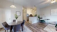 Located at 6700 Cantata St NW in Albuquerque, New Mexico, this one-bedroom, one-bathroom single-floor residence offers a well-designed layout that maximizes every inch of its compact footprint. The open-concept main living area seamlessly integrates the living room, dining room, and kitchen into one cohesive space. Wood-look plank flooring runs throughout the main areas, lending a clean, modern aesthetic, while a ceiling fan adds comfort to the living room. Natural light filters in through windows dressed with grommet-panel curtains, brightening the space throughout the day. The kitchen features a peninsula with a dark granite-style countertop, white cabinetry, stainless steel appliances including a refrigerator, microwave, and range, and track lighting overhead. The dining area accommodates a round table with seating for four beneath a pendant light fixture. The bedroom offers plush carpeting, a comfortable scale, and a dedicated closet. The bathroom is finished with white cabinetry, a dark countertop vanity, a large mirror, wood-look flooring, and a full tub-shower combo. An in-unit laundry area adds everyday convenience. The overall ambiance is light, neutral, and move-in ready.