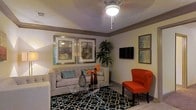 This single-floor space showcases a thoughtfully designed living room that balances comfort and contemporary style. The room is anchored by a tufted gray sofa accented with decorative pillows, complemented by a bold orange accent chair that adds a striking pop of color to the warm, neutral palette. A geometric-patterned area rug in black and white grounds the seating arrangement, while a sleek glass coffee table serves as a functional centerpiece.

Warm ambient lighting is provided by a ceiling fan fixture overhead, casting a cozy glow throughout the room. The walls are adorned with framed artwork in coordinating tones, adding depth and character to the space. A wall-mounted flat-screen television is positioned for comfortable viewing, flanked by additional framed prints.

Crown molding runs along the upper perimeter of the room, adding an architectural finishing touch that elevates the overall aesthetic. A tall, lush indoor plant brings a natural, organic element to the corner of the room, enhancing the inviting atmosphere. Light-toned flooring ties the space together seamlessly. Overall, this living space exudes a warm, curated ambiance that is both functional and visually appealing.