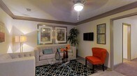 This single-floor space showcases a thoughtfully designed living room that balances comfort and contemporary style. The room is anchored by a tufted gray sofa accented with decorative pillows, complemented by a bold orange accent chair that adds a striking pop of color to the warm, neutral palette. A geometric-patterned area rug in black and white grounds the seating arrangement, while a sleek glass coffee table serves as a functional centerpiece.

Warm ambient lighting is provided by a ceiling fan fixture overhead, casting a cozy glow throughout the room. The walls are adorned with framed artwork in coordinating tones, adding depth and character to the space. A wall-mounted flat-screen television is positioned for comfortable viewing, flanked by additional framed prints.

Crown molding runs along the upper perimeter of the room, adding an architectural finishing touch that elevates the overall aesthetic. A tall, lush indoor plant brings a natural, organic element to the corner of the room, enhancing the inviting atmosphere. Light-toned flooring ties the space together seamlessly. Overall, this living space exudes a warm, curated ambiance that is both functional and visually appealing.