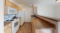 This single-floor residence offers a thoughtfully designed layout with a blend of functional and comfortable living spaces. The living room serves as the centerpiece of the home, featuring a tile-surround fireplace flanked by two windows that invite natural light and outdoor views, all set atop warm, neutral carpeting. A stepped platform adds architectural interest and a sense of depth to the space.

The kitchen is efficiently arranged with light wood cabinetry, dark laminate countertops, recessed lighting, and tile flooring. White appliances including a range, microwave, and refrigerator provide all essential cooking amenities, while an extended countertop offers additional prep and serving space.

The bedrooms are generously proportioned, each carpeted and featuring built-in closet storage. One bedroom includes a mirrored sliding-door closet with multiple shelves and track lighting overhead, while another benefits from a corner window with outdoor views and baseboard heating. A third room provides a double-door closet and wall sconce lighting, offering versatile use as a bedroom or den.

Two full bathrooms mirror each other in finish, both showcasing tile flooring, warm-toned countertops with single-basin vanities, light wood cabinetry, and tub-shower combinations with sliding glass enclosures, providing a clean and cohesive aesthetic throughout.