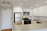 Located at 156 Herchimer Ave in Belleville, Ontario, this single-floor unit offers a well-designed open-concept layout finished with light hardwood flooring throughout. The spacious living area is a standout feature, anchored by a curved floor-to-ceiling window wall that invites abundant natural light and provides elevated views of the surrounding cityscape. Baseboard heating runs along the perimeter, and roller blinds on each window offer adjustable light control.

The kitchen is thoughtfully appointed with white shaker-style cabinetry extending to the ceiling, a white subway tile backsplash, stone-look countertops, and a full suite of stainless steel appliances including a refrigerator, range, microwave, and dishwasher. Recessed pot lighting illuminates the workspace, and a peninsula counter connects the kitchen to the living area.

The bedrooms are comfortably sized, each featuring large windows with roller blinds, hardwood flooring, and access to the hallway. The bathroom is clean and modern, equipped with a sliding glass-enclosed shower, a vanity with storage, and bright overhead lighting. The overall aesthetic is neutral and bright, with white walls and light finishes creating a cohesive, move-in ready environment throughout.