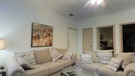 This single-floor residence offers a warm and inviting atmosphere designed for comfortable everyday living. The living room serves as the heart of the home, featuring a pair of plush, neutral-toned sofas adorned with patterned accent pillows that add visual interest without overwhelming the space. A glass-topped coffee table anchors the seating area, while a large framed landscape painting in warm earth tones adds a sophisticated artistic touch to the wall. The room is finished with smooth, light-colored walls and tiled flooring that reflects light and contributes to a clean, airy feel. Overhead, a ceiling-mounted light fixture provides ample illumination throughout the space. Two interior doors visible from the living area lead to additional rooms, suggesting a functional and well-connected floor plan. The overall design aesthetic leans toward a relaxed, neutral palette, creating a cohesive and comfortable environment. The layout of the living space allows for easy movement and versatile furniture arrangement, making it both practical and welcoming for residents and guests alike.