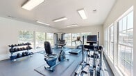 This fitness center in Southaven, Mississippi is a well-equipped, single-level facility designed to support a well-rounded workout routine. The main exercise area is bright and welcoming, featuring crisp white walls, durable rubber-matted flooring, and large windows along multiple walls that flood the space with natural light and offer views of the surrounding outdoor areas, including a nearby pool and landscaped grounds. Recessed fluorescent panel lighting supplements the natural light, ensuring excellent visibility throughout the day and evening. The cardio equipment lineup includes True-brand treadmills, elliptical trainers, and recumbent bikes, all positioned to take advantage of the scenic window views. A multi-station cable machine provides options for a variety of strength-training exercises, while a three-tier dumbbell rack stocked with a range of free weights supports traditional lifting routines. A dedicated functional training rack along one wall holds medicine balls, resistance bands, stability balls, foam rollers, and additional accessories, making the space versatile for diverse fitness needs. The facility also includes a bathroom and convenient exterior door access. The overall ambiance is clean, modern, and functional, offering a focused and motivating environment for residents.